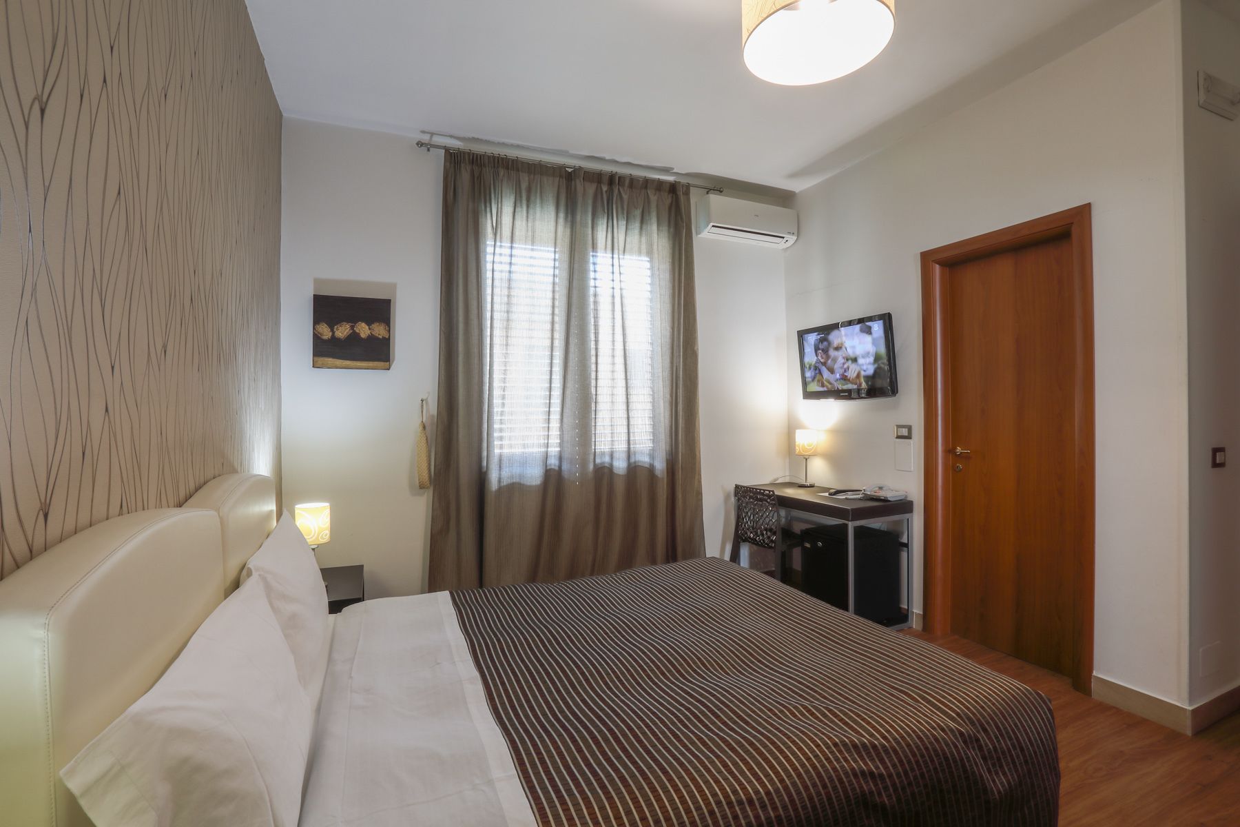A hotel room with a bed, a desk with a TV, a window with curtains, and an air conditioning unit.