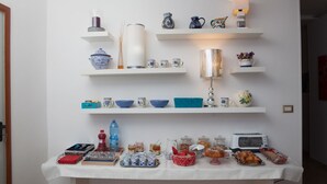 Montaje de desayuno buffet con diferentes alimentos y utensilios de cocina sobre una mesa, y repisas superiores que muestran objetos decorativos y utensilios.