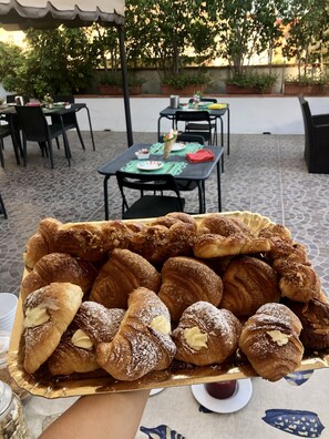 A tray of assorted pastries on a table in an outdoor dining area with chairs and tables, surrounded by potted plants and a tiled floor. - Night and Day B&B (Reggio Calabria)