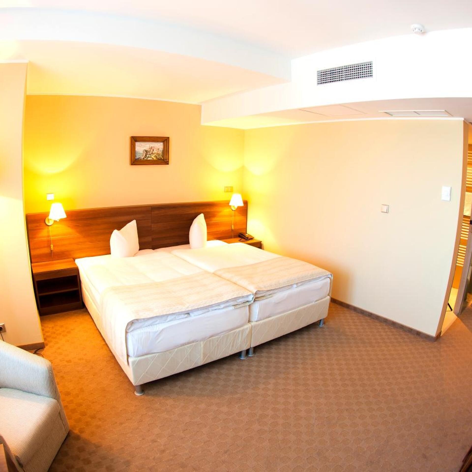 A hotel room with a double bed, two bedside lamps, a headboard, a picture on the wall, and a carpeted floor.