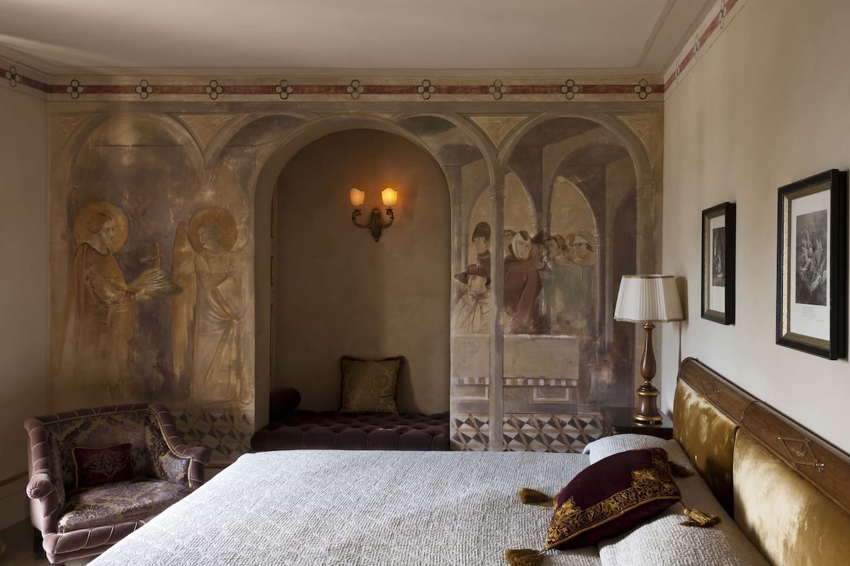 A bedroom with a large bed, a chair, a lamp, and a wall with frescoes.