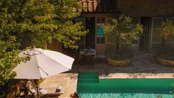 A swimming pool with a tiled deck, surrounded by greenery and a building with a tiled roof.