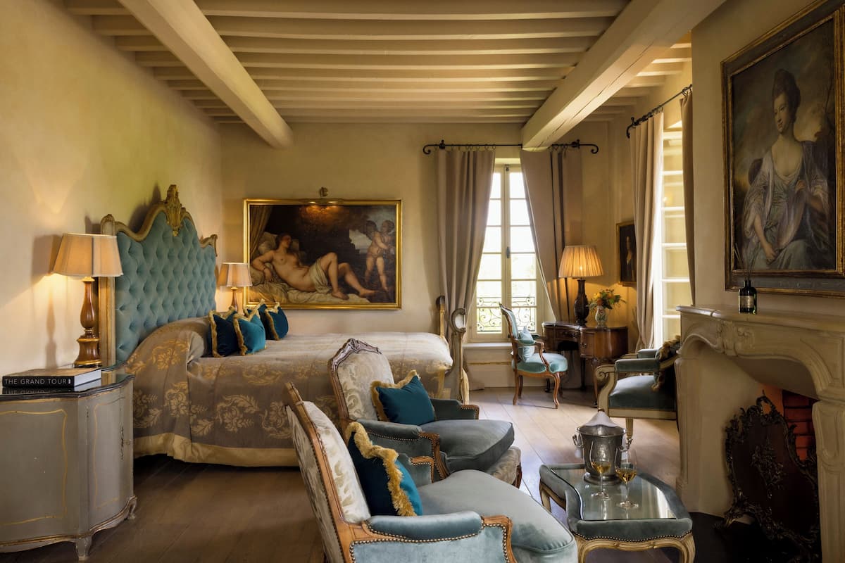 A luxurious bedroom with a large bed, two armchairs, a chaise lounge, a small table with a lamp, and a fireplace.