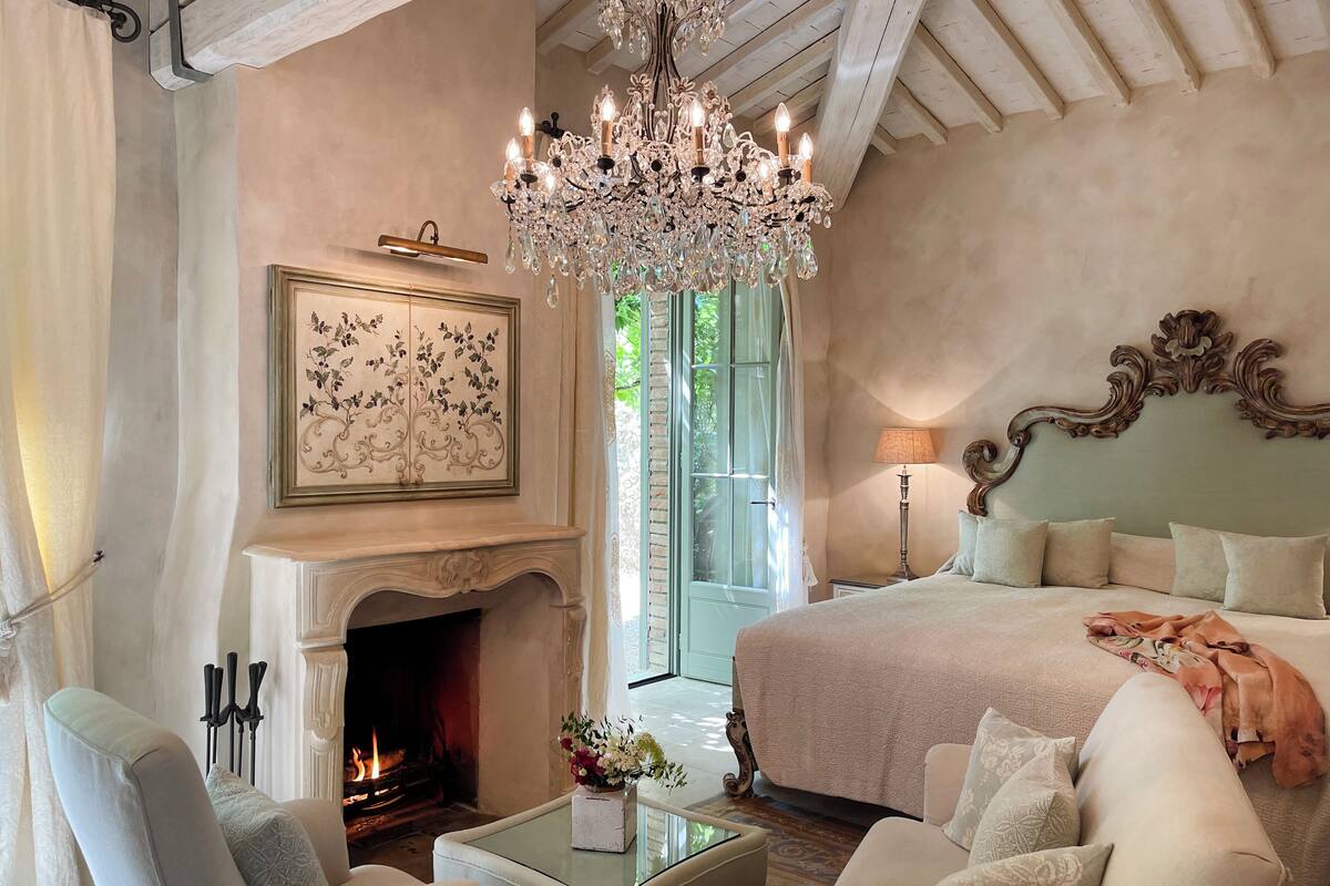 A bedroom with a fireplace, a chandelier, a bed, and a view of greenery outside.
