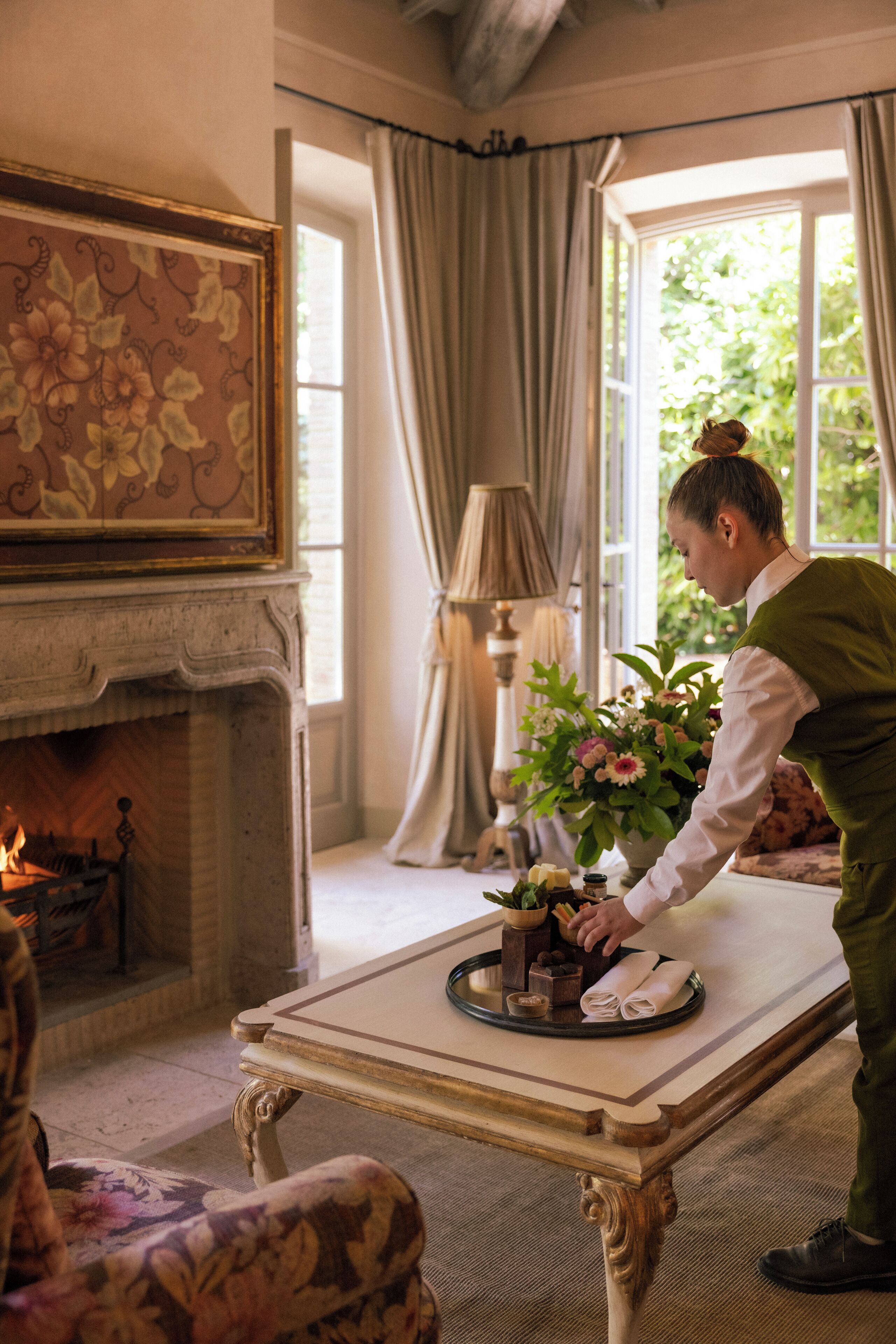 A room with a fireplace, a dining table with a tray of food, a potted plant, and a window with curtains.