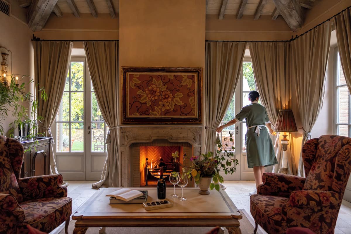 A room with a fireplace, floral armchairs, a table with a book and wine glasses, and a woman standing by the window.