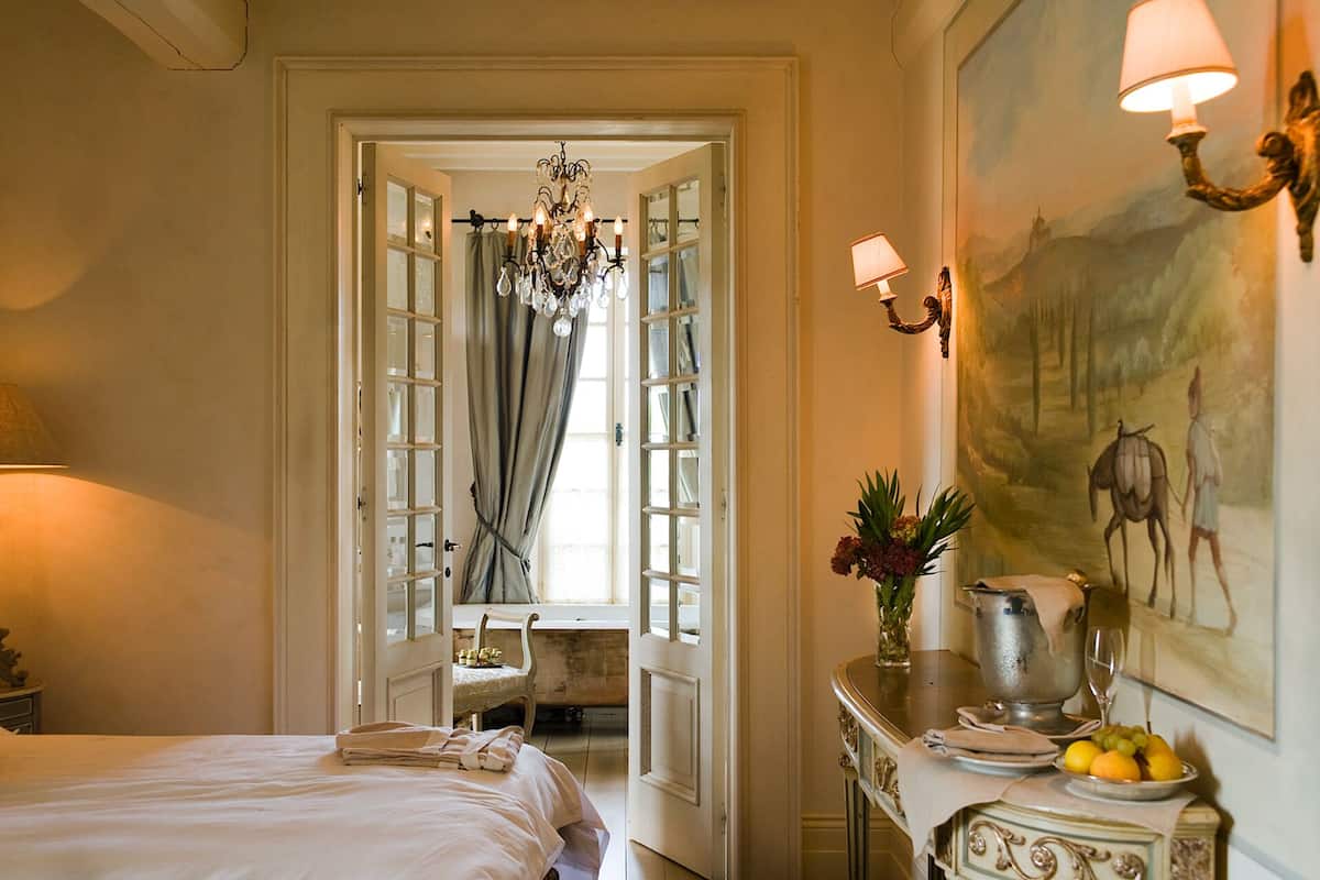 A hotel room with a bed, a chandelier, a painting, a table with a pitcher and fruit, and a chair.