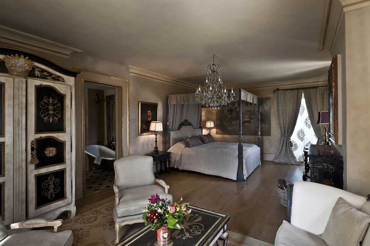 A spacious bedroom with a large bed, a sitting area with a sofa and armchairs, a chandelier, and a classic decor style.