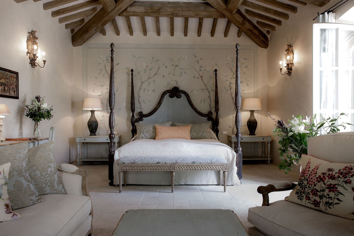 A four-poster bed with a floral headboard, a sitting bench, and a chaise lounge in a room with wooden beams on the ceiling.