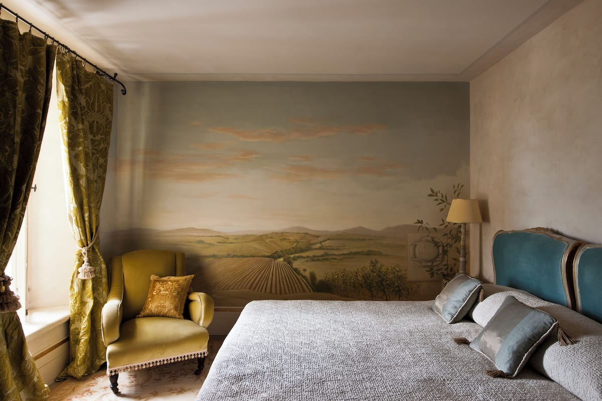 A bedroom with a bed, a chair, a lamp, and a mural on the wall.