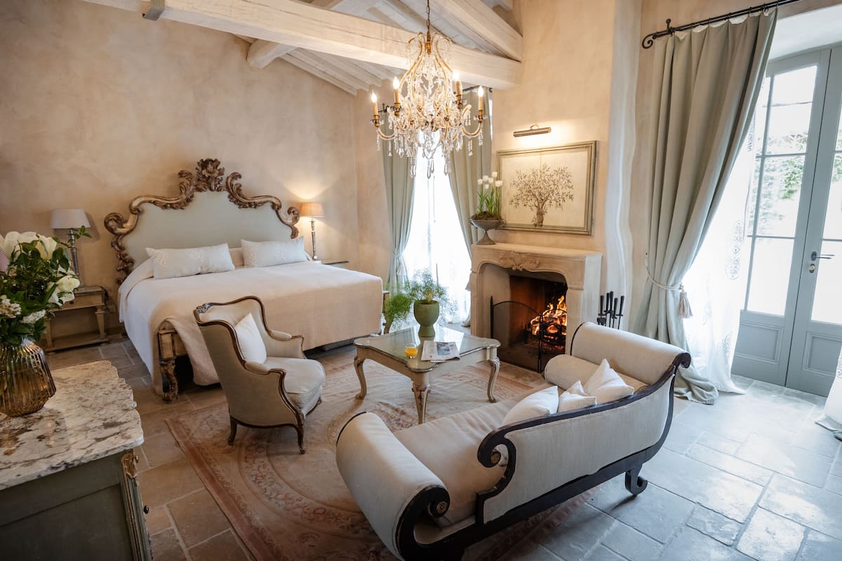 A spacious bedroom with a large bed, two armchairs, a small table, and a fireplace.
