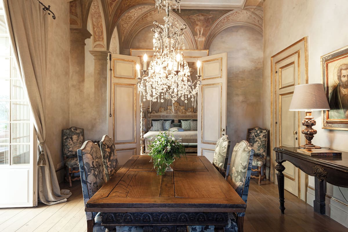 A dining room with a wooden table, ornate chairs, a chandelier, and a bed in the background.