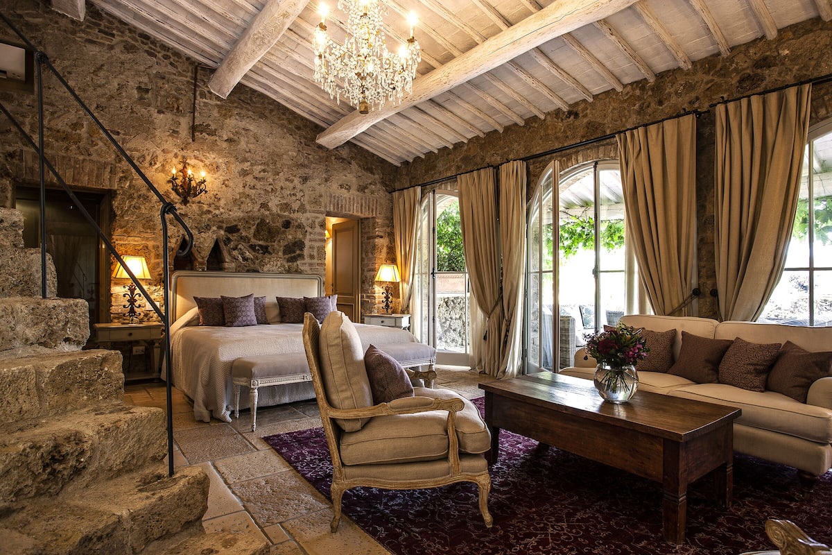 A rustic bedroom with a stone wall, a bed, a chair, a sofa, and a chandelier.