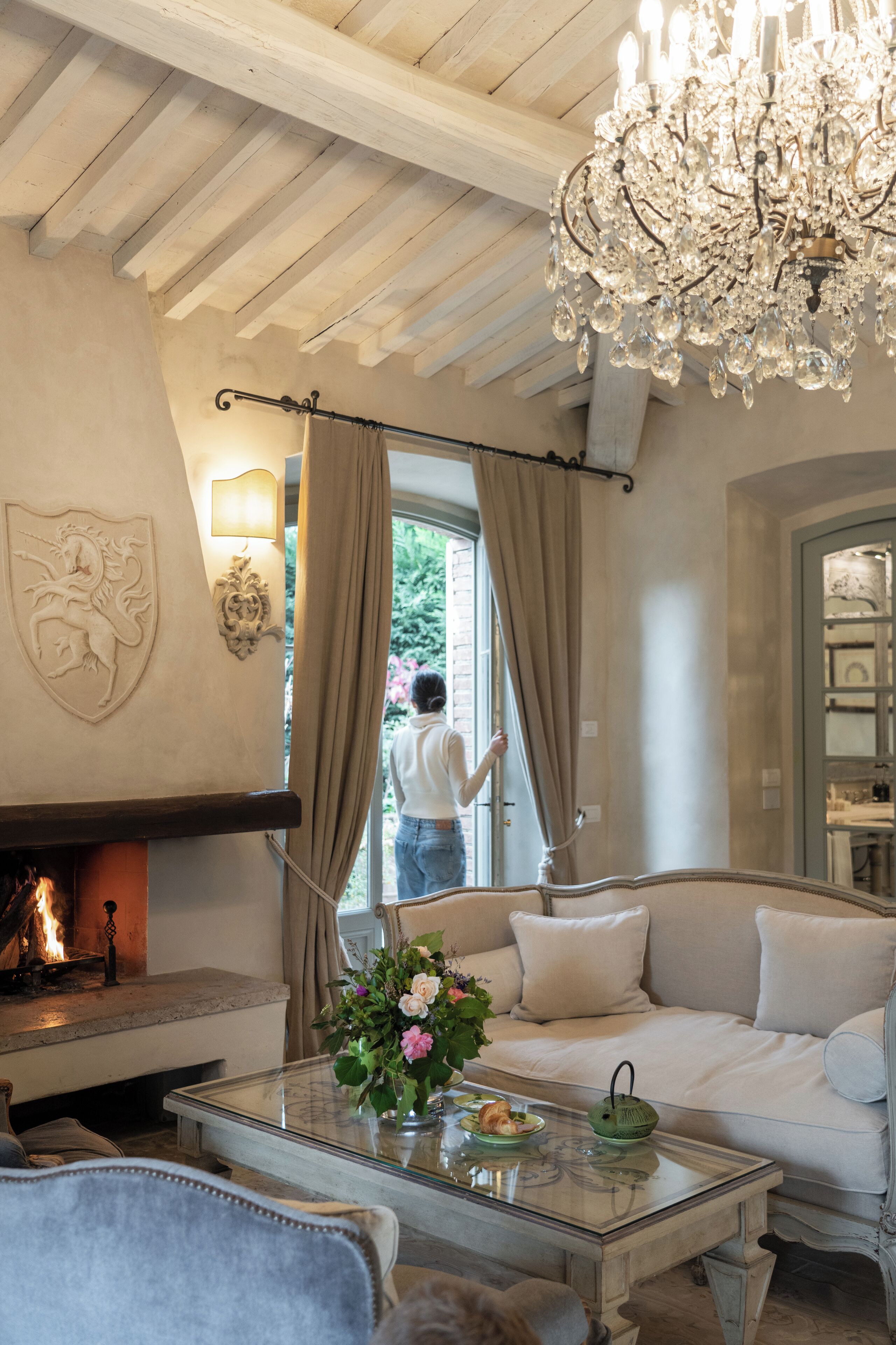 A cozy living room with a fireplace, a sofa, a glass coffee table with flowers, and a chandelier.