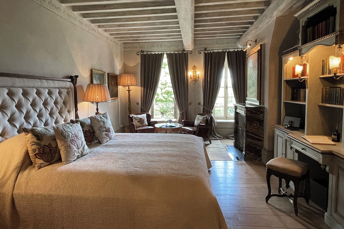 A bedroom with a large bed, two chairs, a desk with a lamp, and a bookshelf.