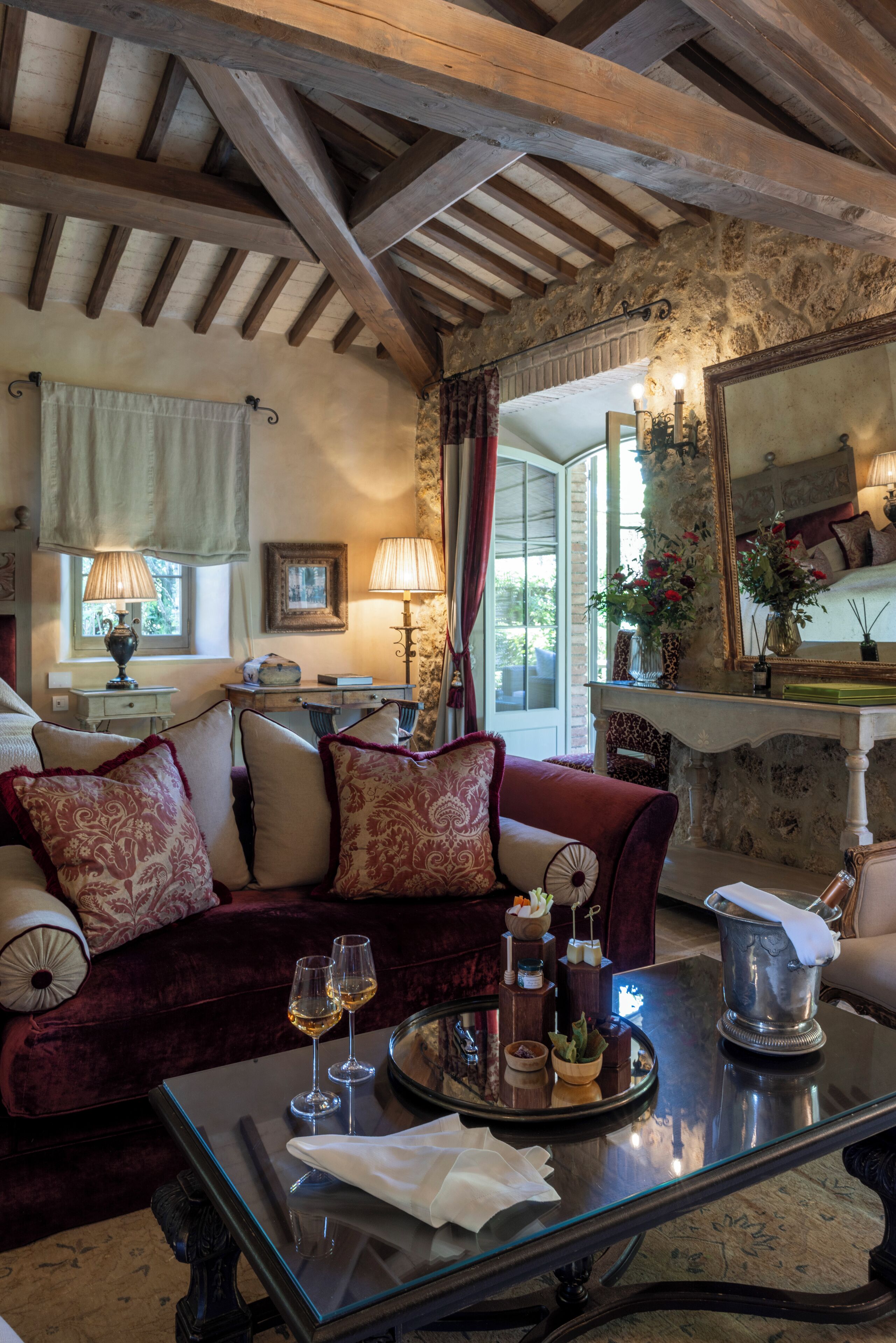 A cozy room with a wooden ceiling, a plush red sofa, a glass coffee table with wine glasses, and a stone wall with a mirror.