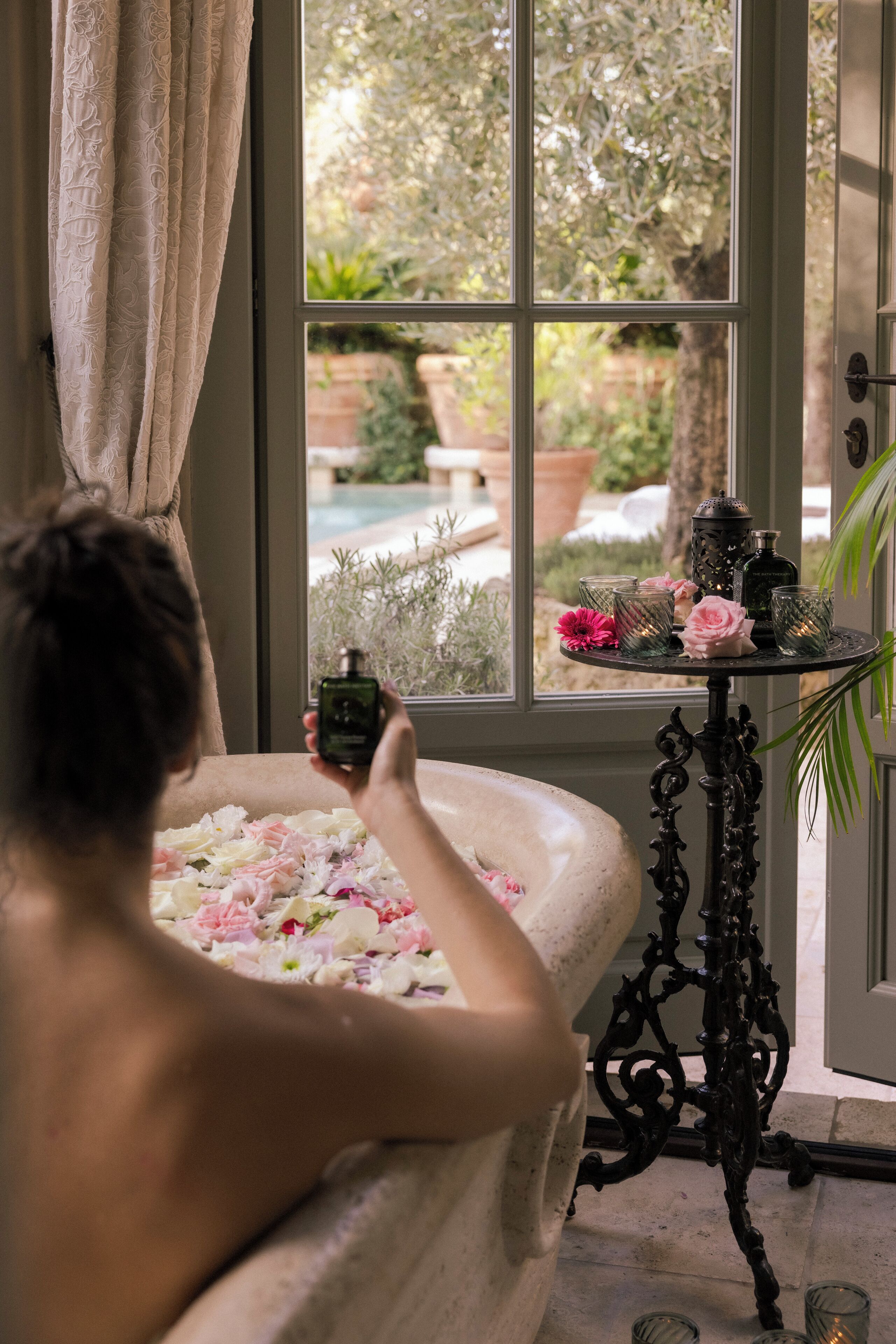 Una vasca da bagno piena di petali di fiori, una boccetta di profumo e un tavolo con candele e un vaso di fiori vicino a una finestra.