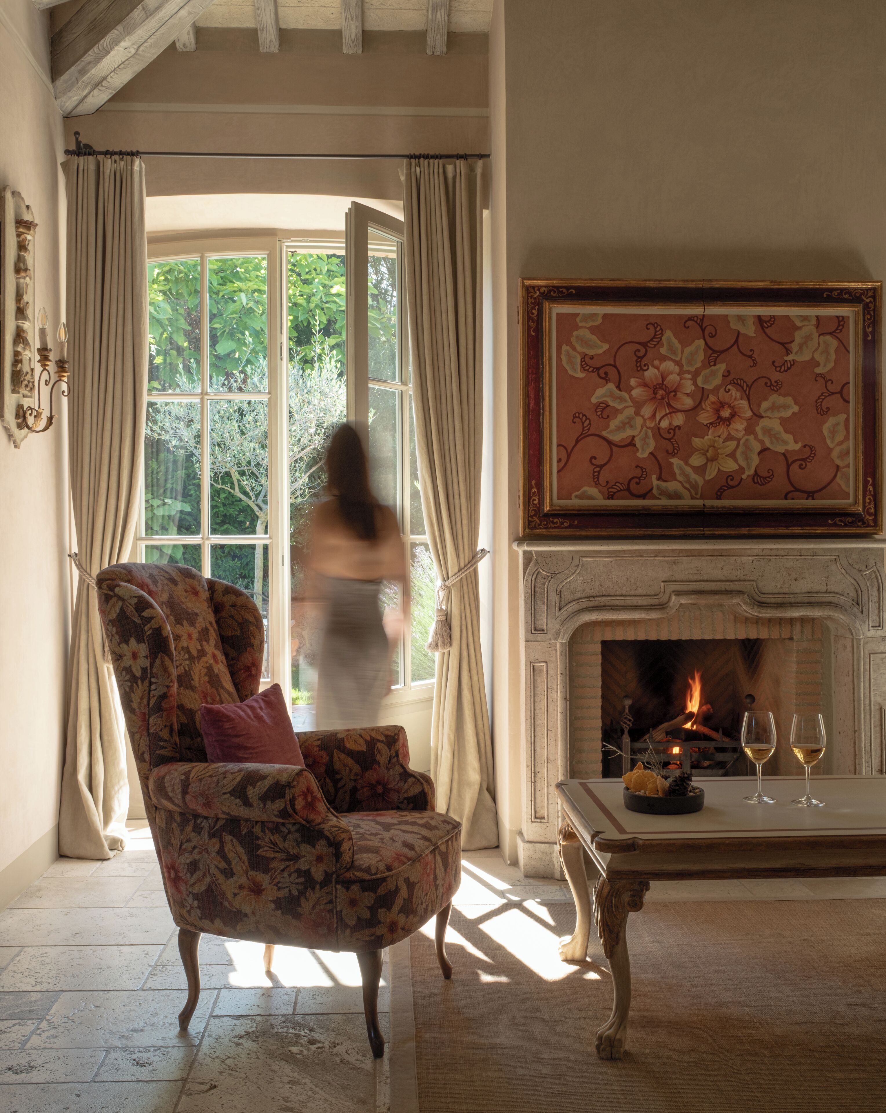 A room with a large window, a fireplace, and a floral armchair.