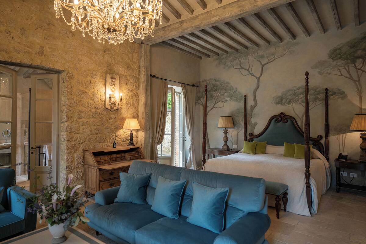 A bedroom with a four-poster bed, a chandelier, stone walls, and a large window.