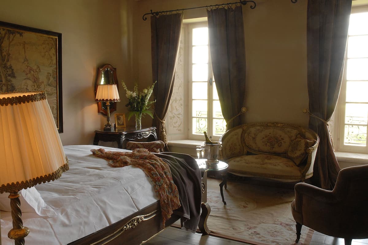 A bedroom with a bed, a chair, a lamp, a vase with flowers, and a painting on the wall.