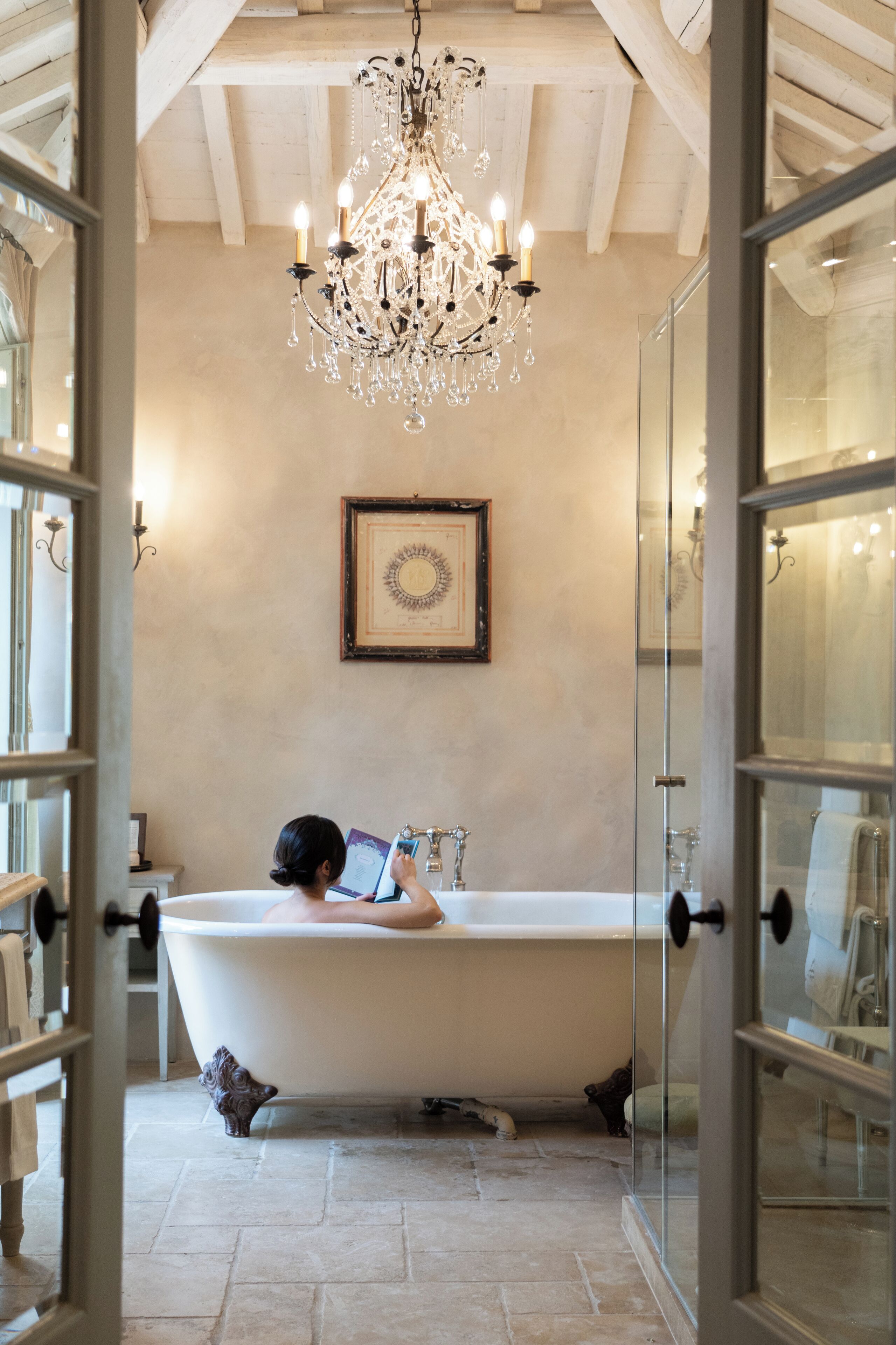 A bathtub in a room with a chandelier, framed artwork on the wall, and a glass door leading to another room.