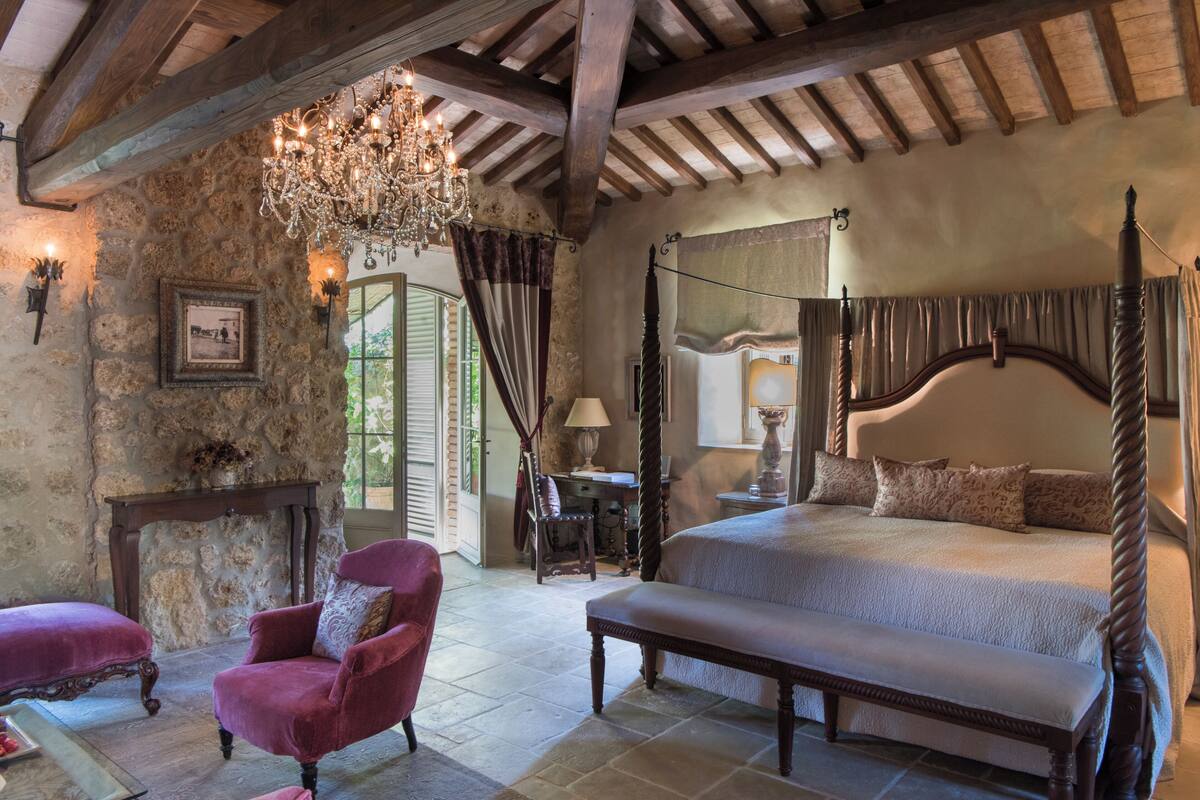 A bedroom with a four-poster bed, a chandelier, a stone wall, and a red armchair.