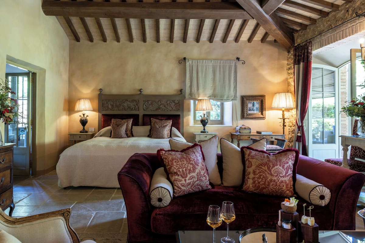 A bedroom with a large bed, two bedside tables with lamps, a red velvet sofa, and a view of the outdoors.