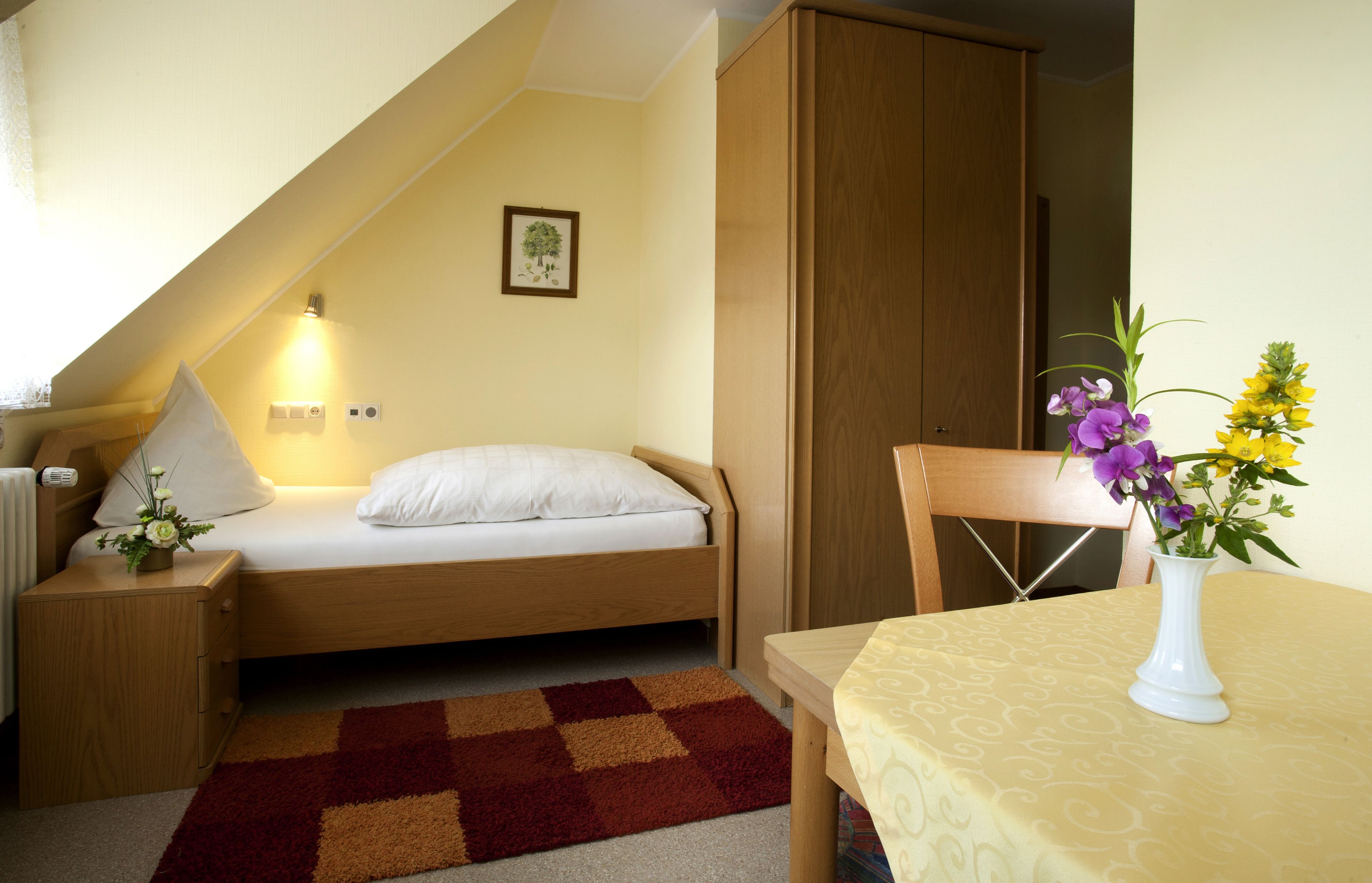 Una camera da letto accogliente e piccola con un letto singolo, un comodino, un tavolino con un vaso di fiori, un'armadio in legno e un piccolo tavolo con tovaglia e un vaso di fiori.