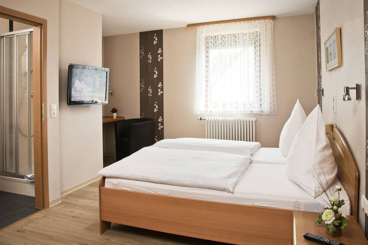 Ein ordentlich eingerichtetes Hotelzimmer mit Bett, Schreibtisch, Stuhl, Fernseher und einem Fenster mit Vorhängen.