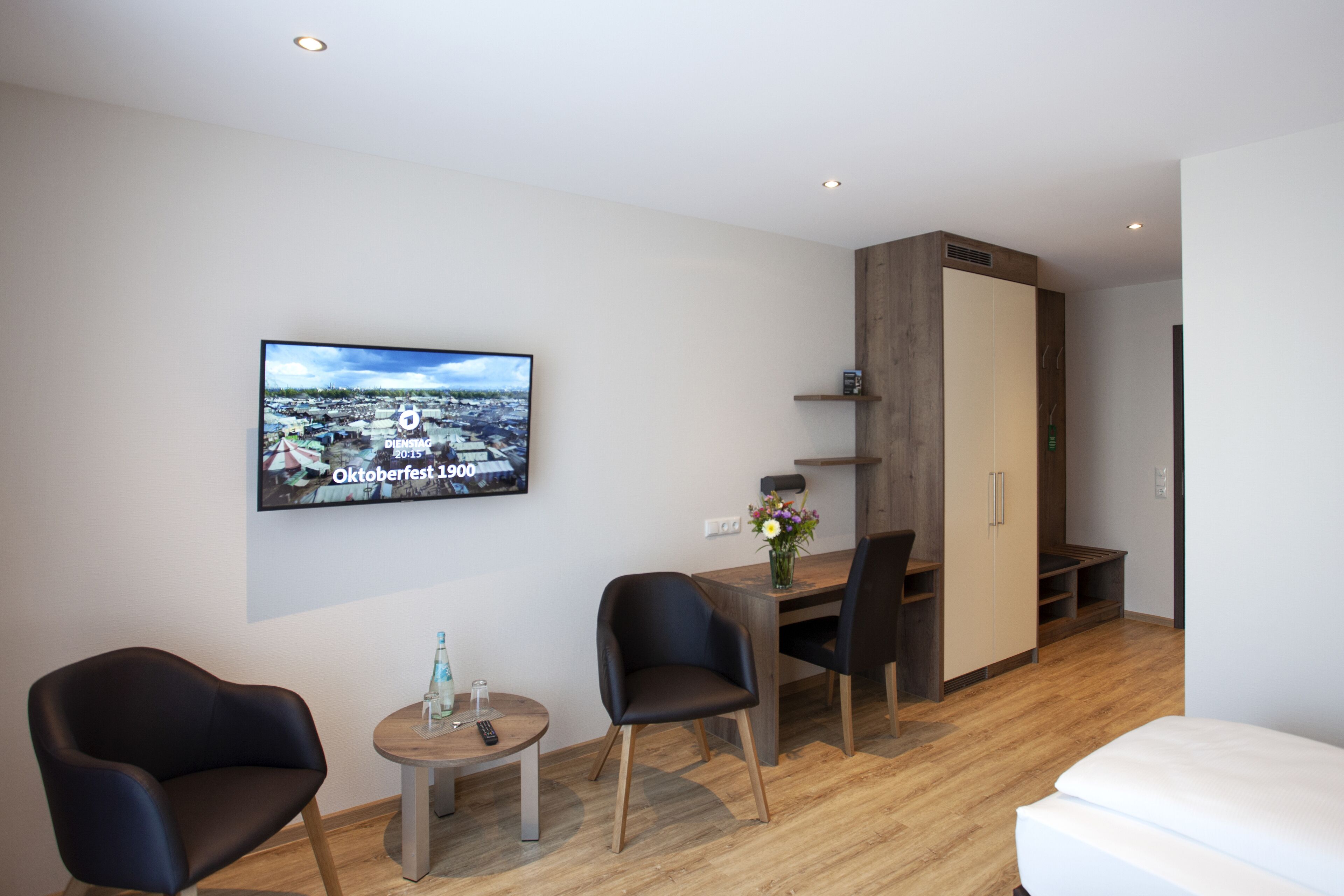 Ein modernes Hotelzimmer mit Holzboden, einem Bett, einem Schreibtisch mit Stuhl, einem an der Wand befestigten Fernseher und einem Kleiderschrank.