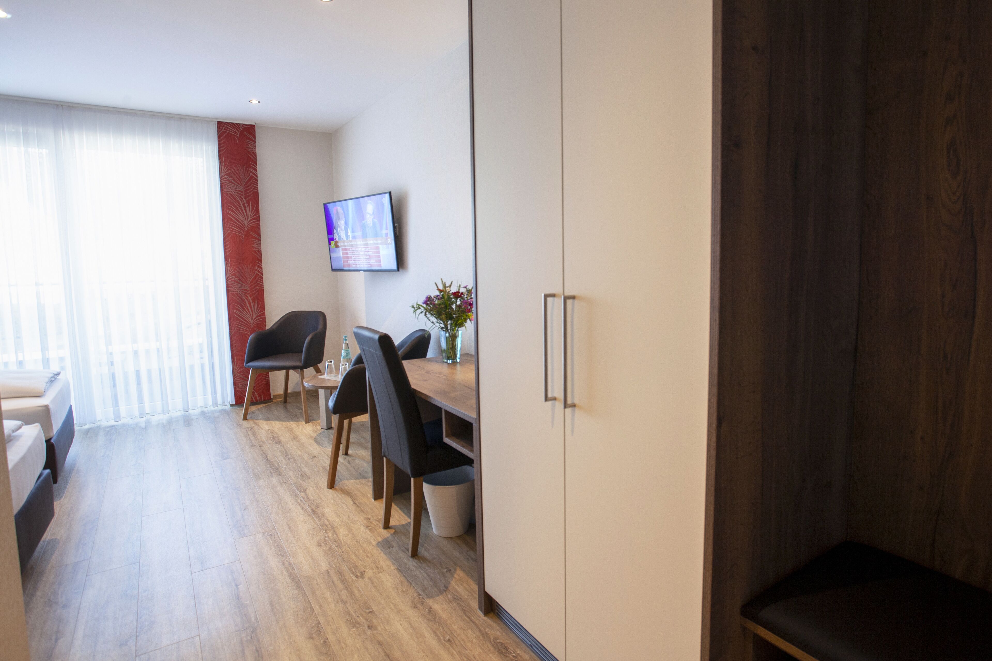 A modern hotel room with a wooden floor, a dining area with a table and chairs, a TV mounted on the wall, and a large wardrobe.