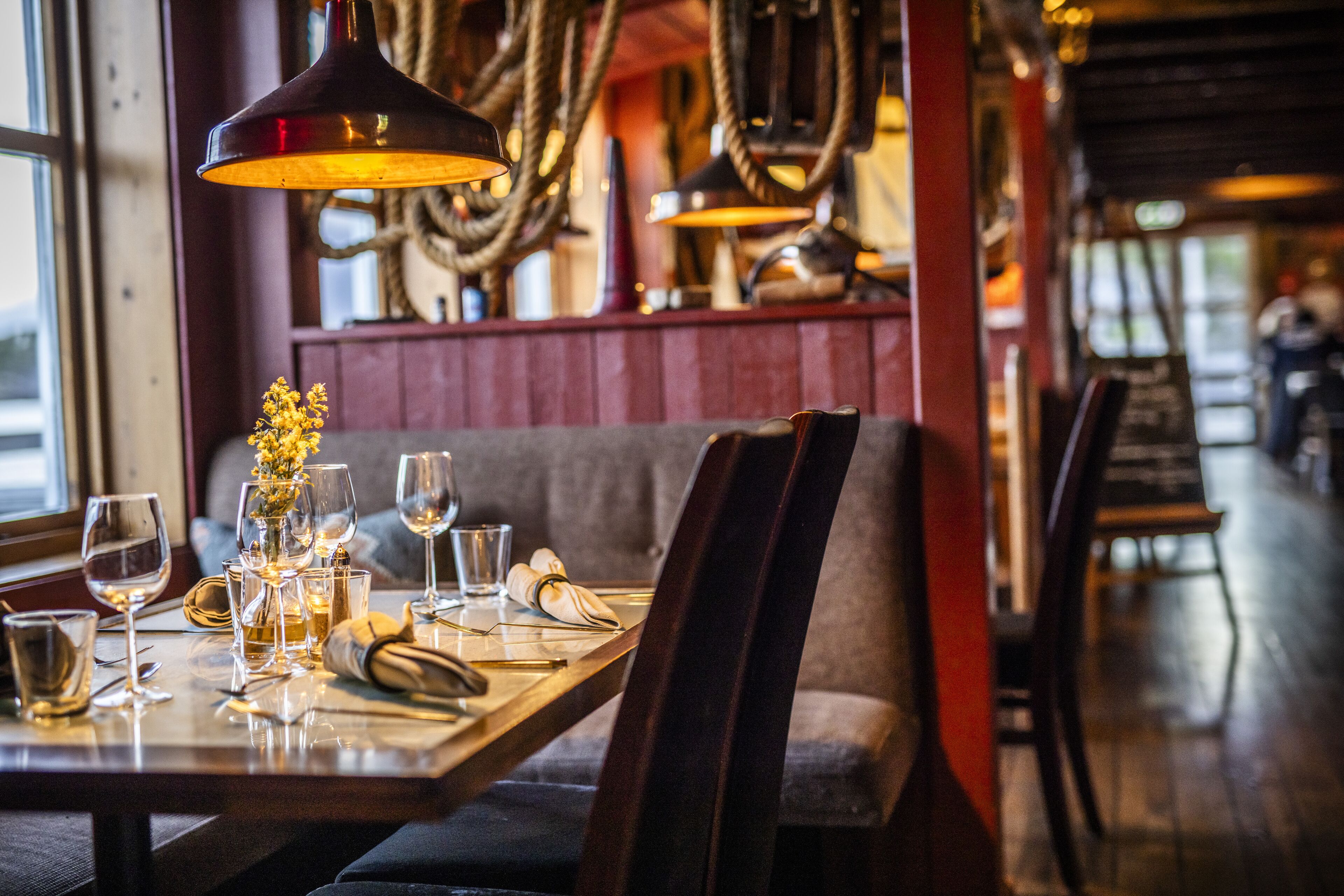 A restaurant interior with a wooden table set for dining, a vase with flowers, and hanging rope lights.