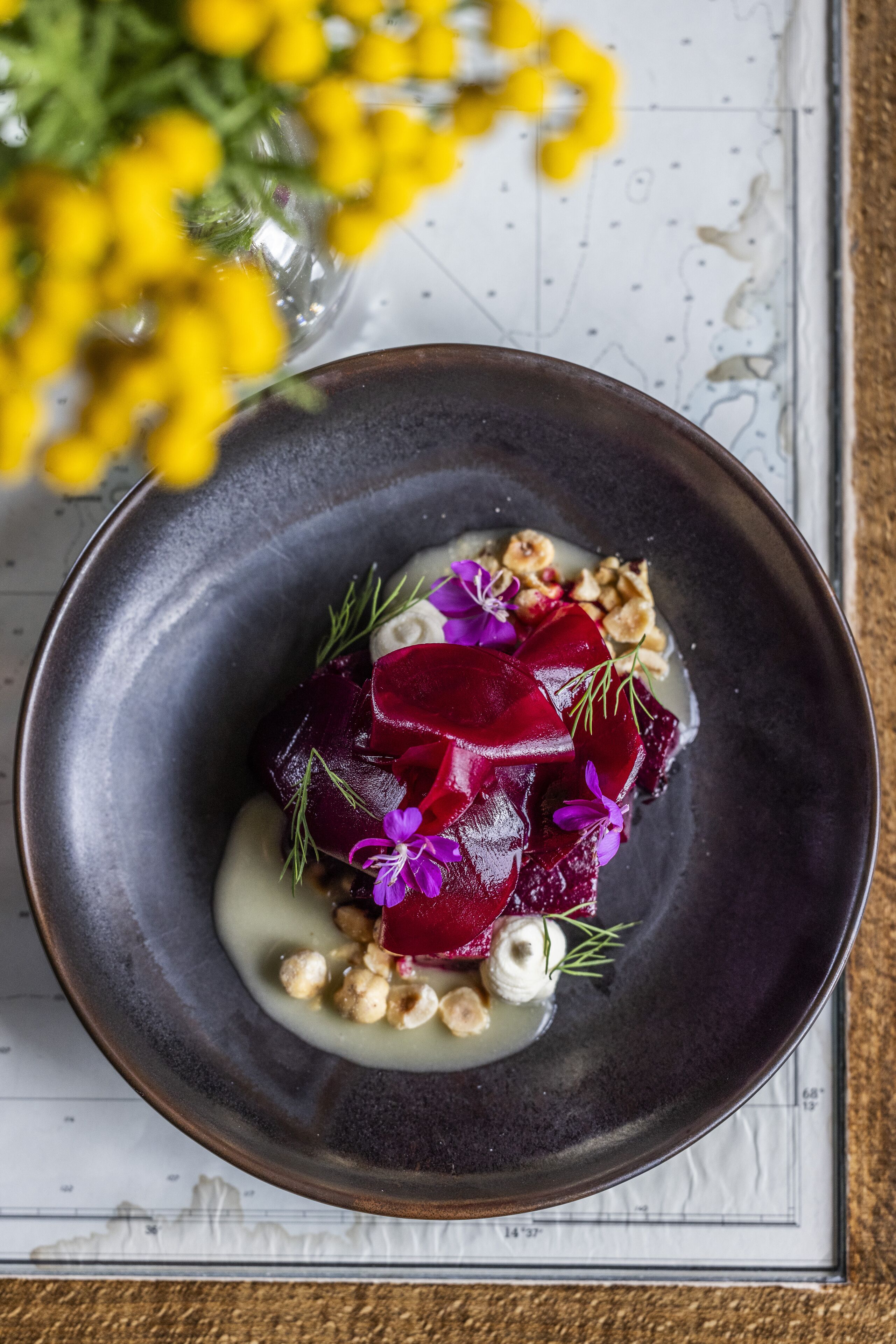 A dark ceramic plate with a beetroot salad, nuts, and flowers on a white surface with a map.