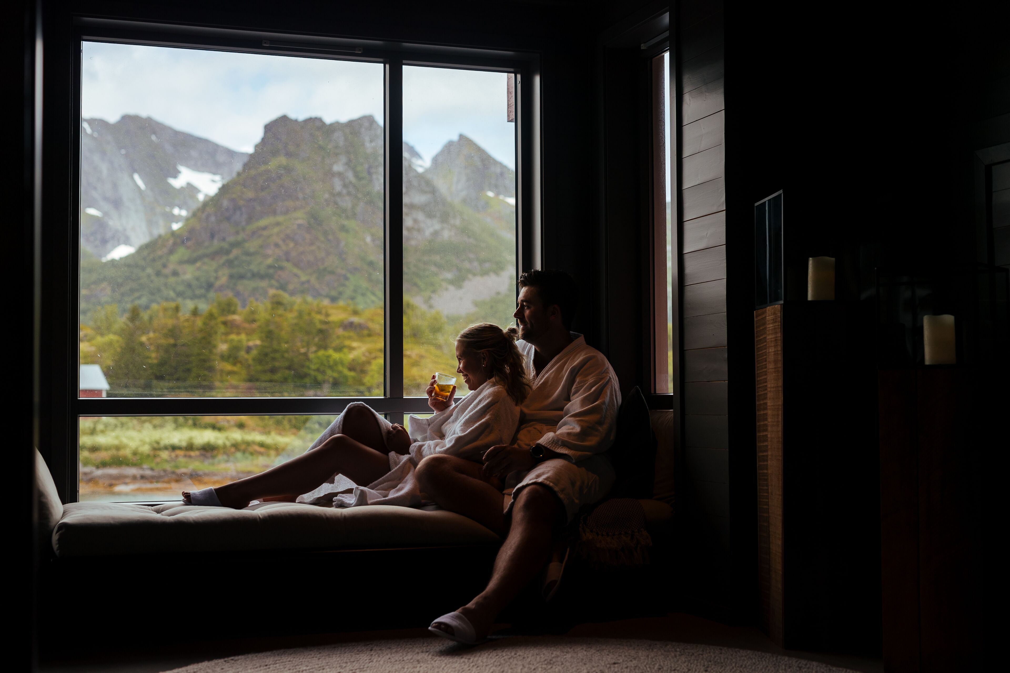 Ein Paar in einem schwach beleuchteten Raum mit einem großen Fenster mit Blick auf eine gebirgige Landschaft.