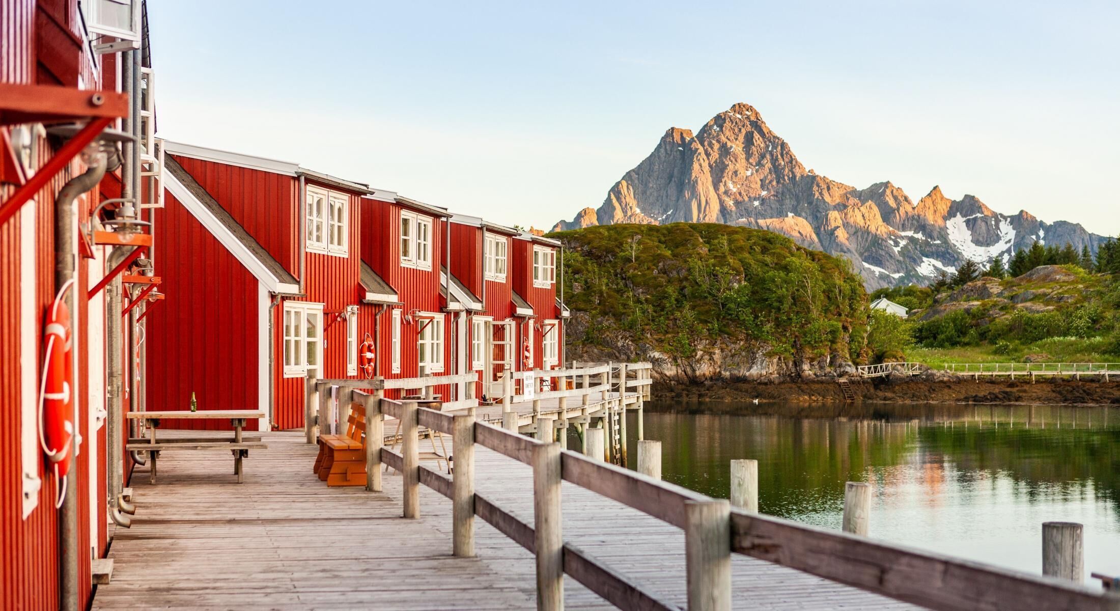 Een rood houten gebouw met een houten terras dat uitsteekt over stil water, met een bergketen op de achtergrond.