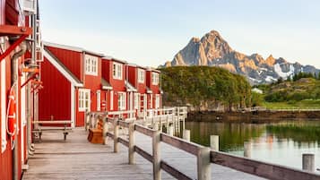 A red wooden building with a wooden deck extending over a calm body of water, with a mountain range in the background.