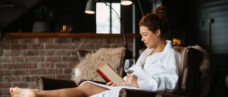 A person relaxing in a leather armchair with a book and glass of wine, in a room with brick walls and modern lighting.