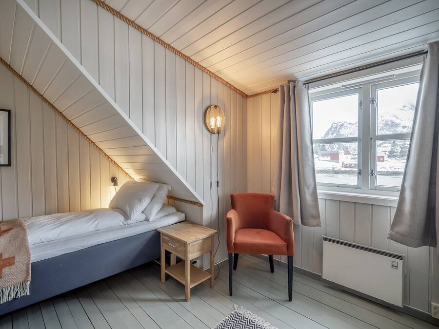 Un dormitorio con cama, una silla, una mesita, una lámpara de pared, una ventana con cortinas y un radiador.