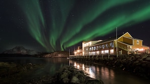 Un edificio de madera con una terraza que se extiende sobre un cuerpo de agua, bajo un cielo nocturno con estrellas visibles y auroras boreales.