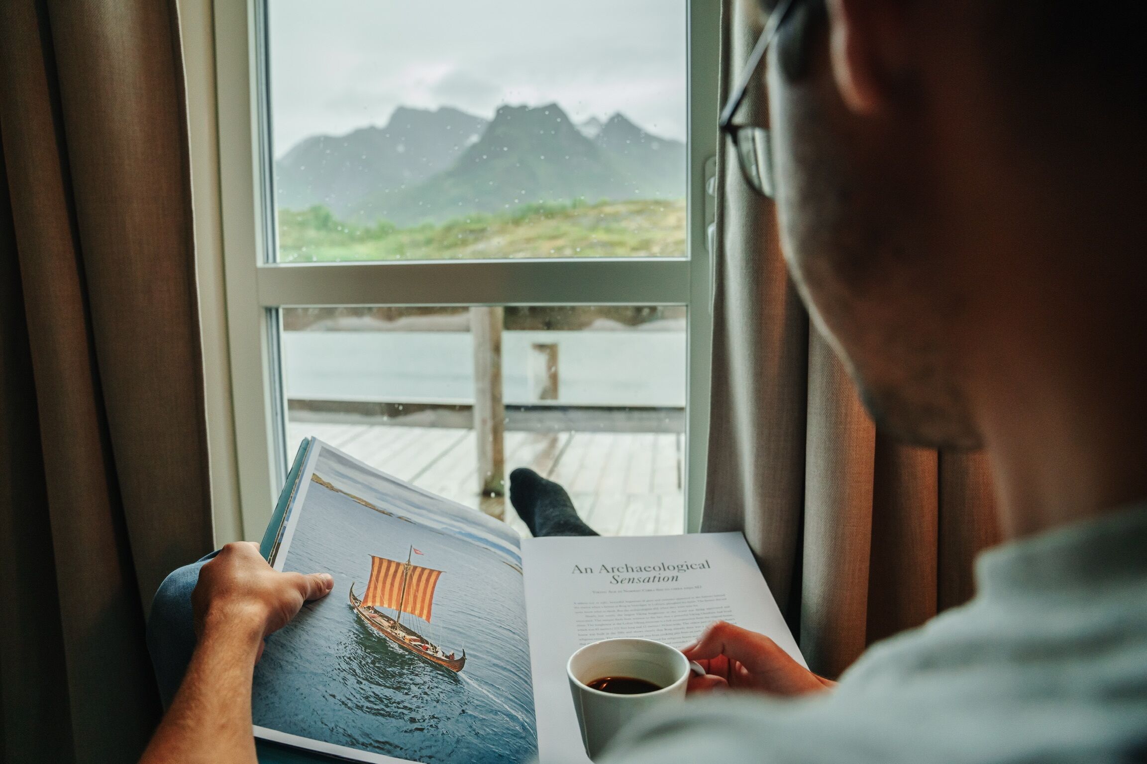 A person reading a book by a window with a view of mountains and a body of water.