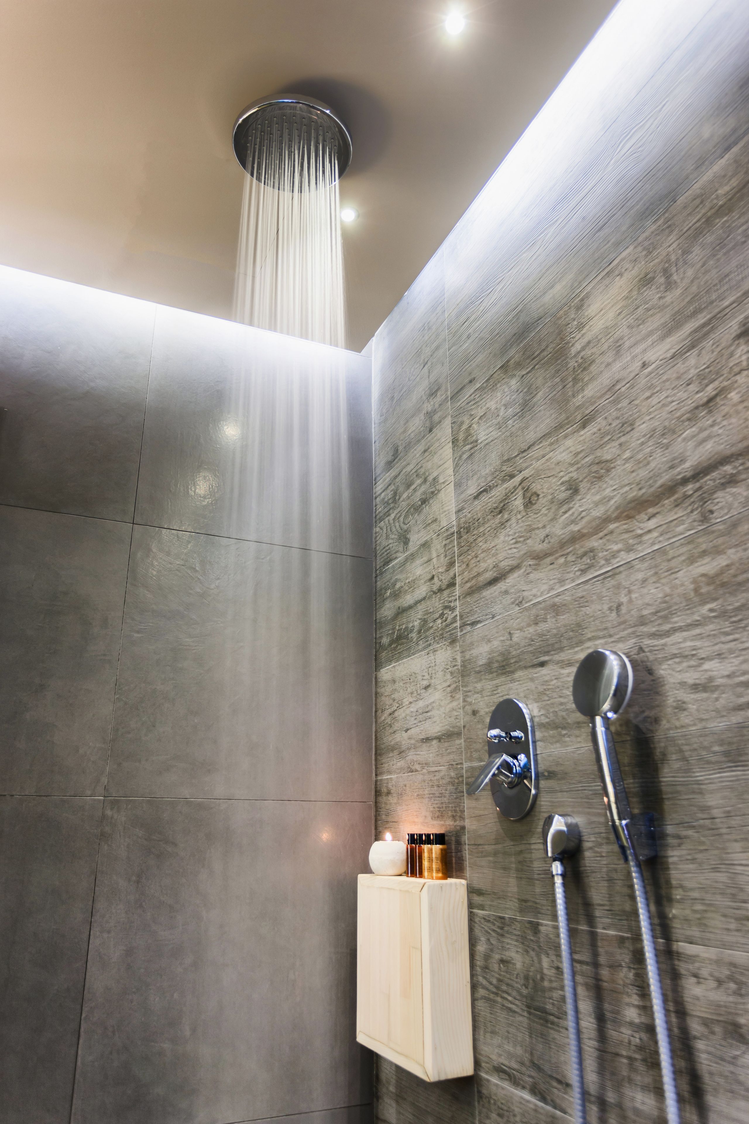 A modern shower with a rain showerhead, a handheld showerhead, and a soap dispenser on a wooden panel wall.
