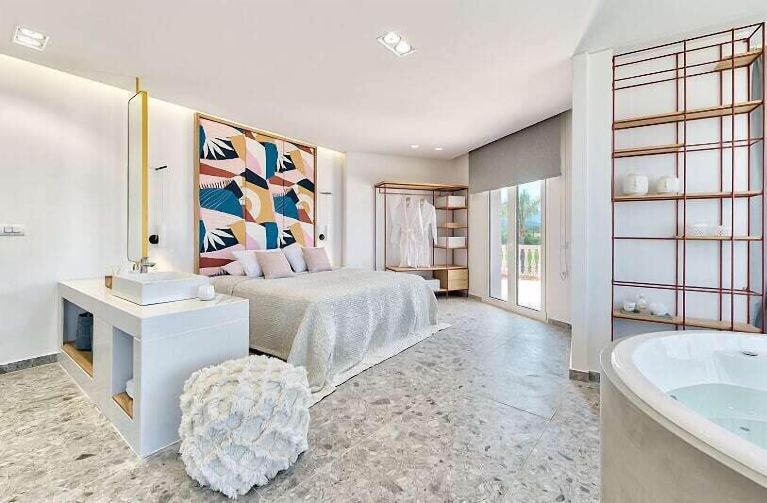 A modern bedroom with a large bed, a white vanity, a bathtub, and a large window.