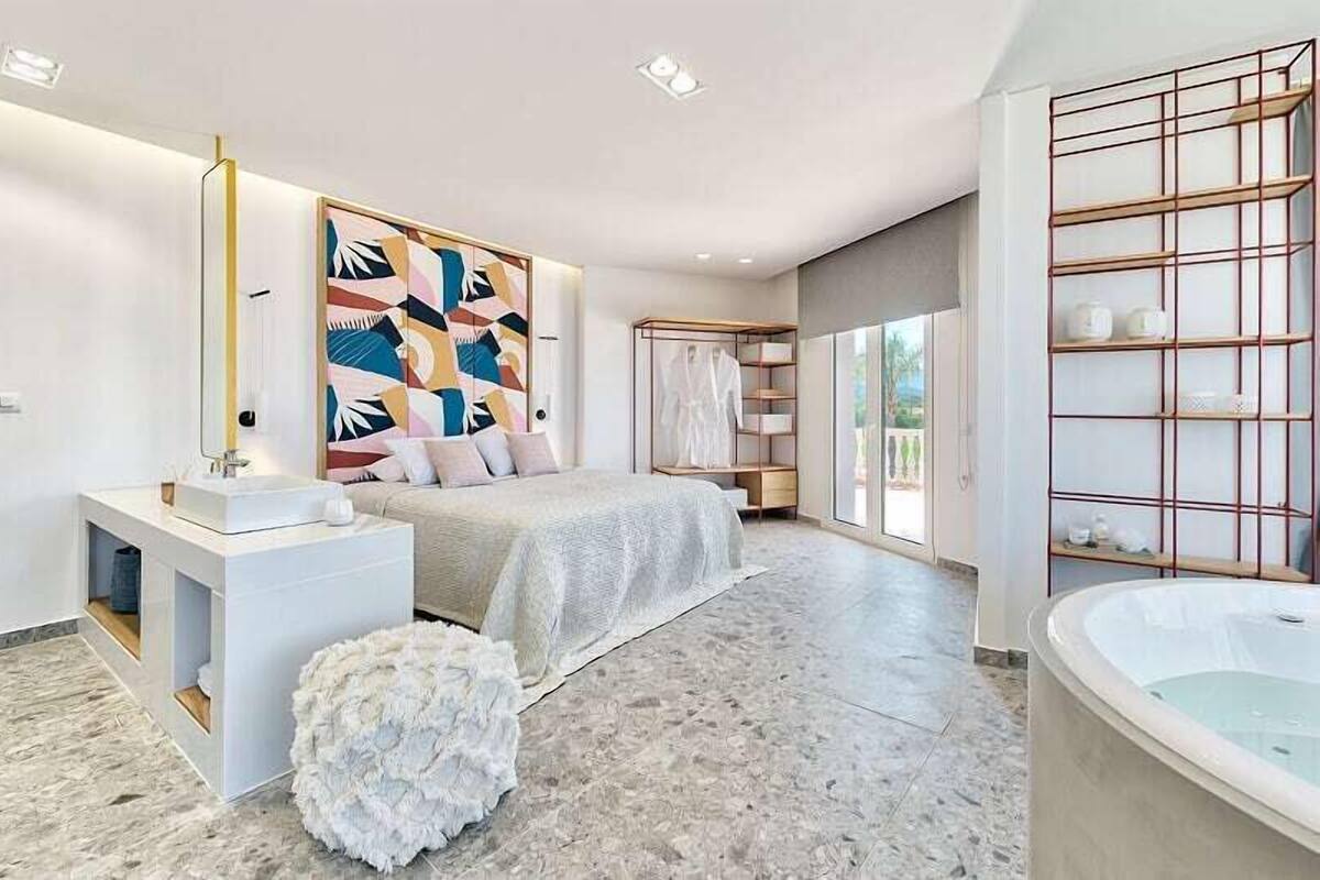 Un dormitorio moderno con una cama grande, un tocador blanco, una bañera y un ventanal amplio.