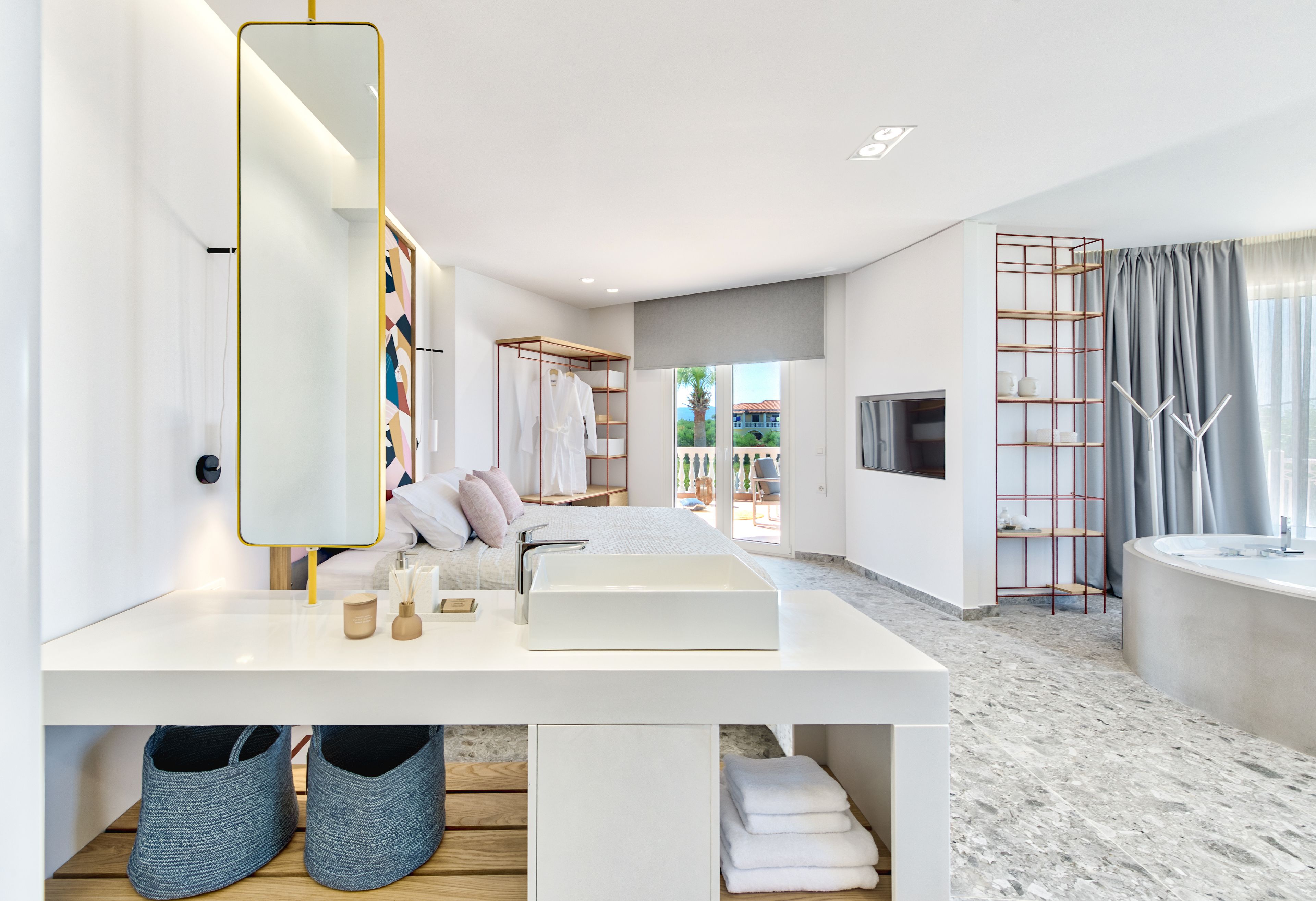 A modern hotel room with a white vanity, a large mirror, a bed with white linens, a TV mounted on the wall, and a bathroom with a large sink and a shower.