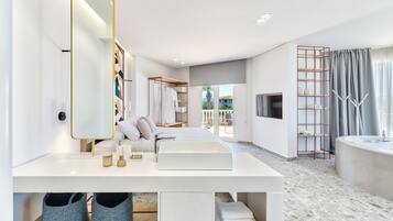 A modern hotel room with a white vanity, a large mirror, a bed with white linens, a TV mounted on the wall, and a bathroom with a large sink and a shower.