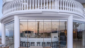 A bar area with a curved white balustrade, glass walls, wicker furniture, and a view of the landscape. - Arkadia Luxury Hotel Apartments (Zakynthos)