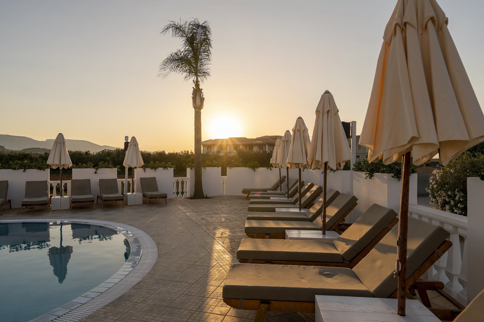A poolside area with lounge chairs and umbrellas, a palm tree, and a sunset in the background.