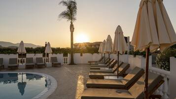A poolside area with lounge chairs and umbrellas, a palm tree, and a sunset in the background.