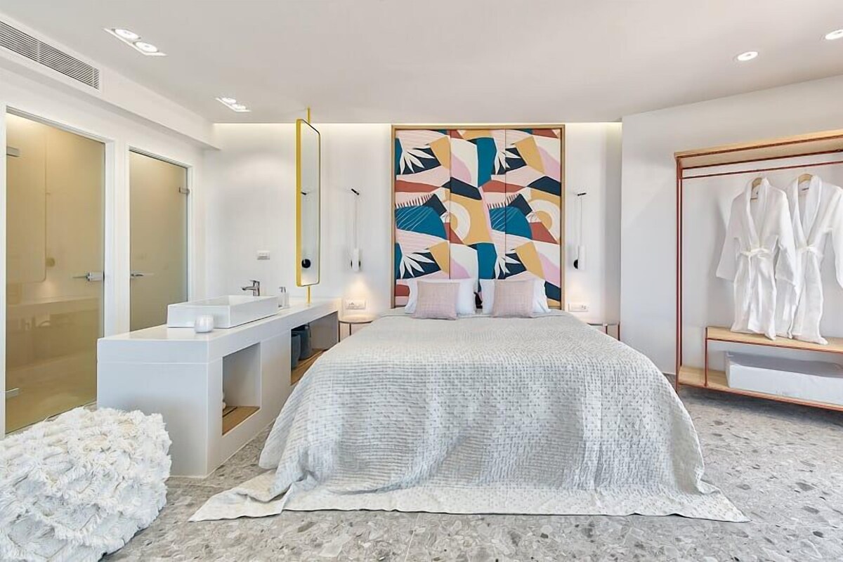 A modern hotel room with a large bed, a vanity with a mirror, a robe rack, and a shower area.