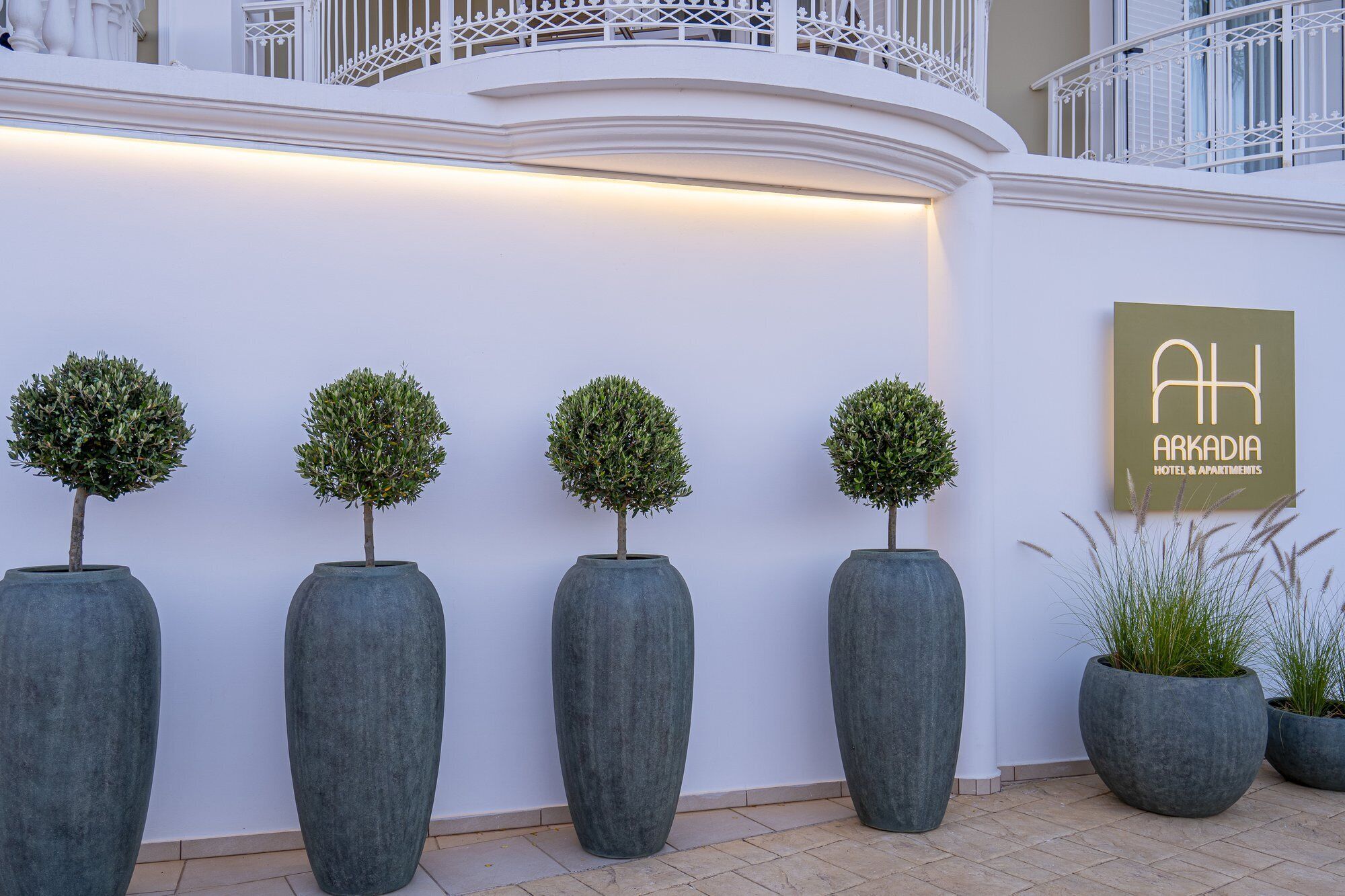 Une rangée de quatre grands bacs à fleurs gris, surmontés d’arbres taillés en forme de sphère, une enseigne indiquant « AH ARKADIA HOTEL & APARTMENTS », et une balustrade blanche.